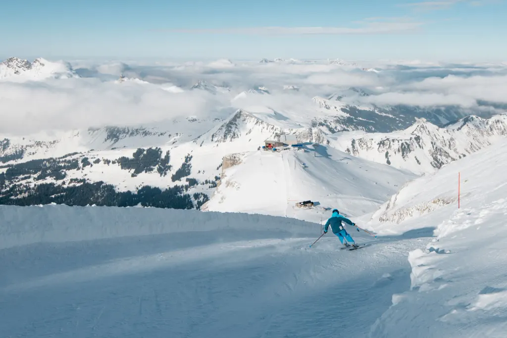 Narciarz na trasie lodowca Glacier 3000, poniżej stacja wyciągu i szczyty Alp Vaud nad morzem chmur