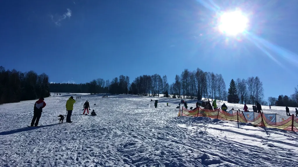 Szeroki ośnieżony stok ośrodka Samoty koło Železnej Rudy w słońcu, z narciarzami i lasem w tle