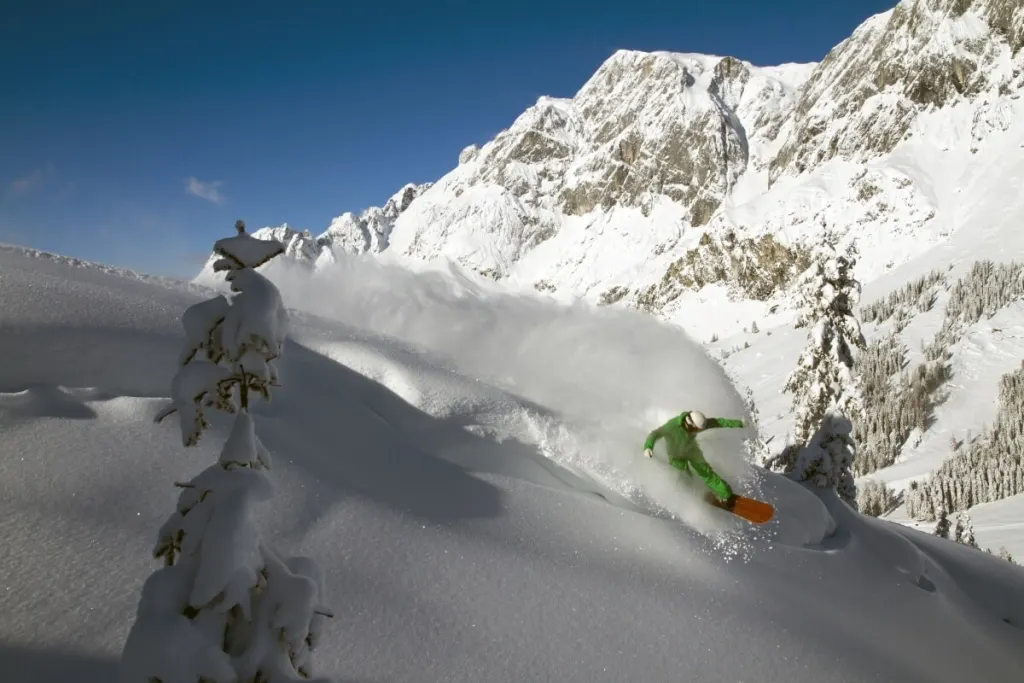 Snowboardzista w głębokim puchu na Hochkeil, nad nim skalny masyw Hochkönig w słońcu