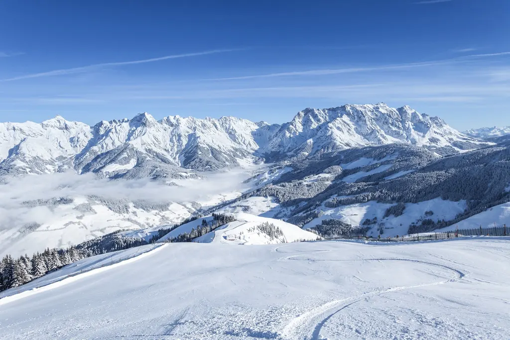 Wyratrakowane trasy ośrodka Hochkönig zimą, w tle masyw Hochkönig nad morzem chmur w dolinie