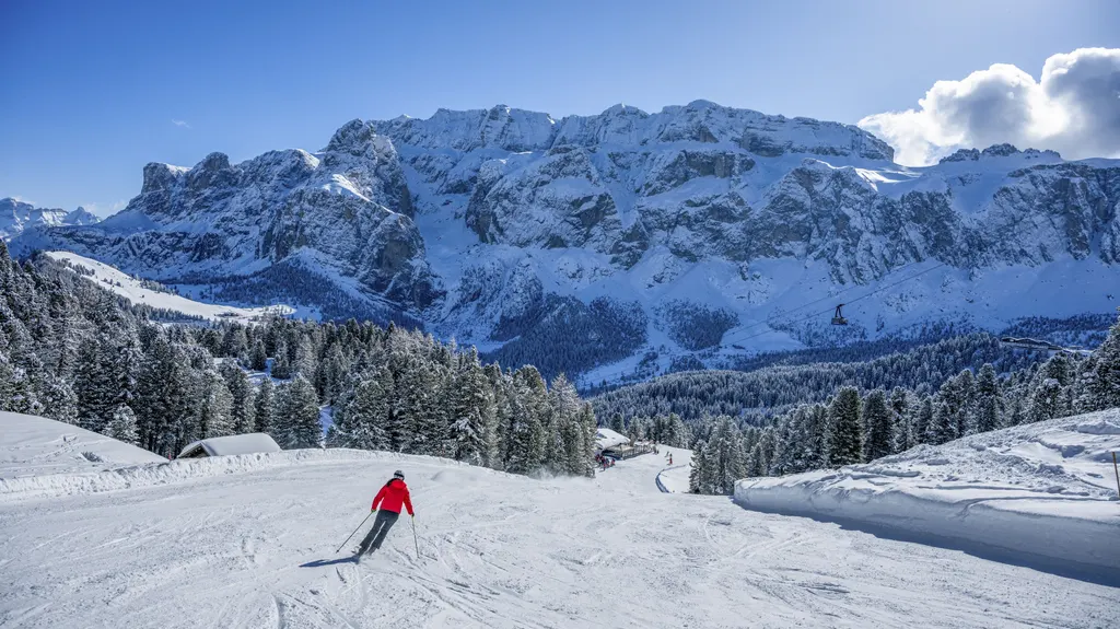 Narciarz na szerokiej trasie w Val Gardena, w tle ośnieżony masyw Sella i zalesione zbocza doliny